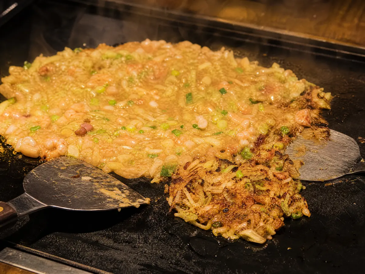 Loose monjayaki batter spread on a hot iron griddle, one portion scraped and caramelising at the edge