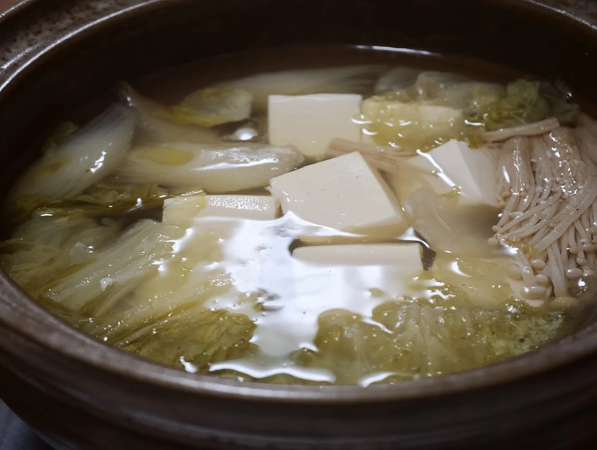 An earthenware pot of yudofu simmering over a flame with dipping sauce and garnishes