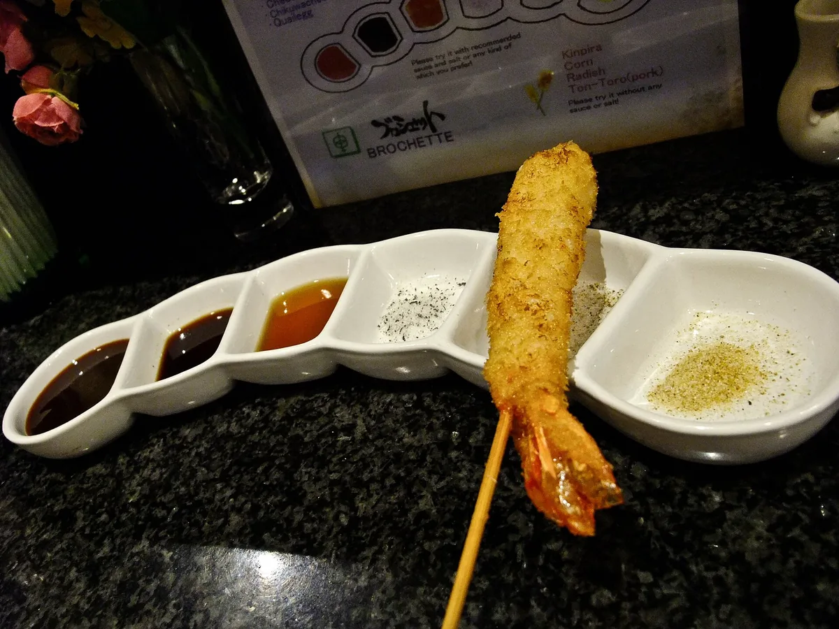 Kushikatsu skewers beside a shared stainless sauce tray on a counter