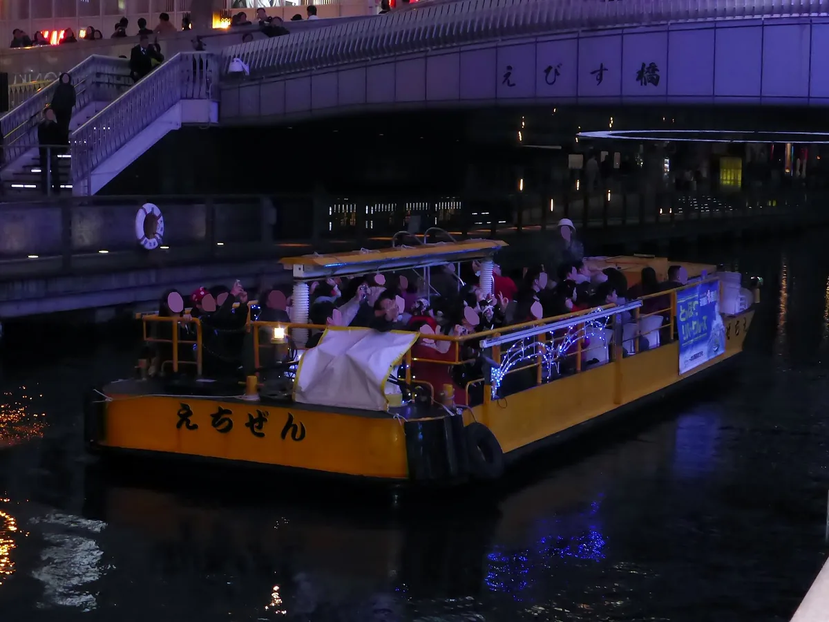 An open sightseeing boat passing under neon signs on the Dotonbori canal