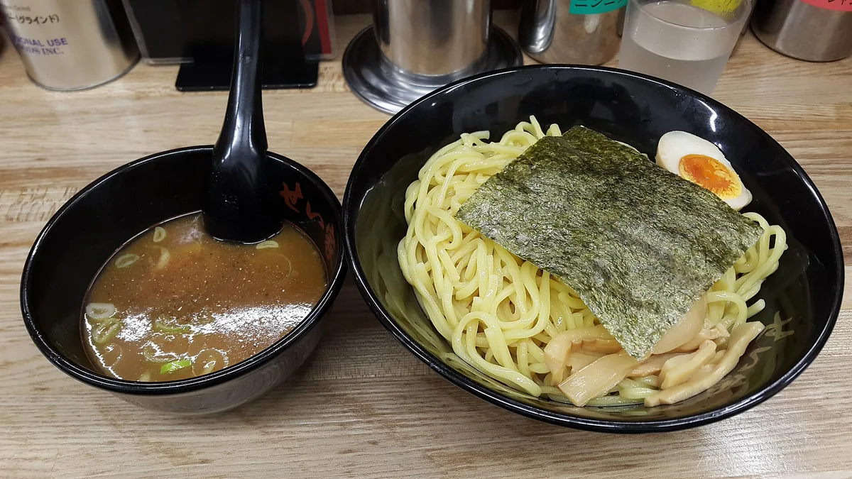 A bowl of thick cold noodles beside a small bowl of dark dipping broth